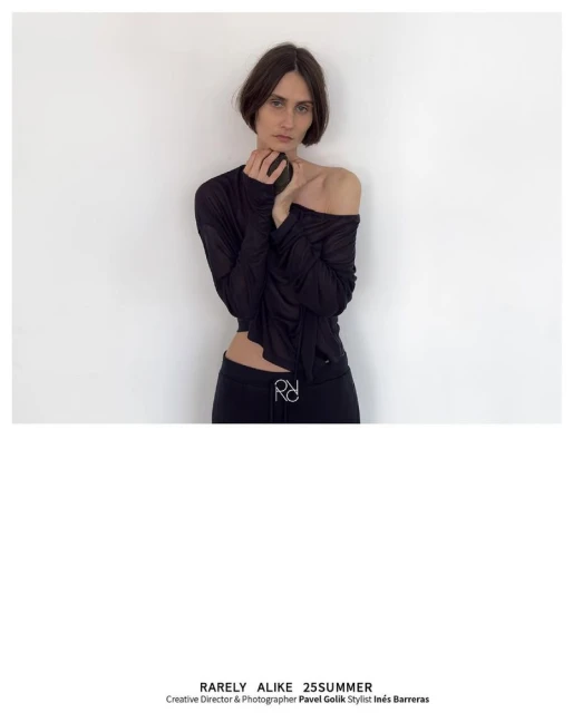 A woman with dark hair is posing in a black top and pants against a white wall in a fashion editorial studio setting.