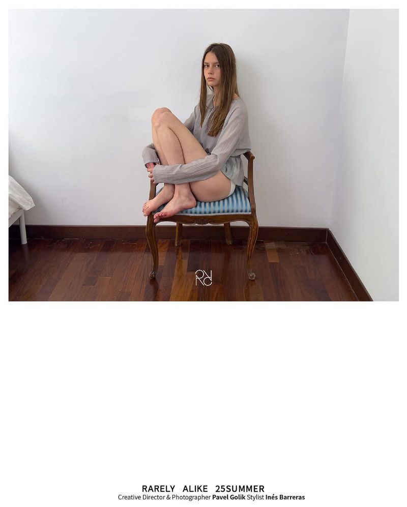 A young woman with long hair sits cross-legged in a chair with a wooden frame. She wears a light-colored top in a minimalist indoor setting.