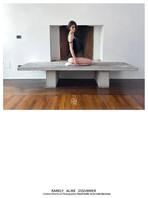 Young woman in black leotard poses on concrete bench in bright room with parquet floor and white walls, minimalist design.