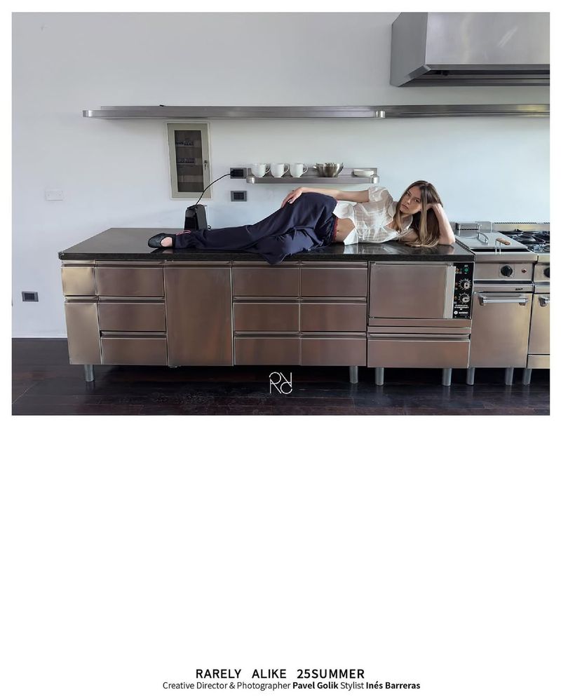 Fashion shot of a woman reclining on a stainless steel kitchen counter, wearing a cropped white top and blue pants.