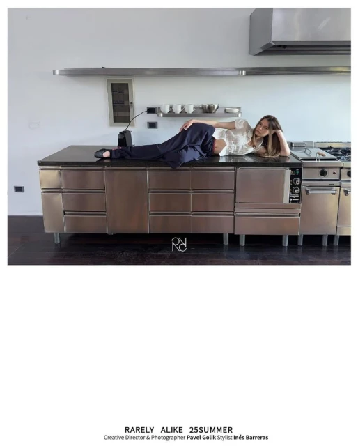 Fashion shot of a woman reclining on a stainless steel kitchen counter, wearing a cropped white top and blue pants.