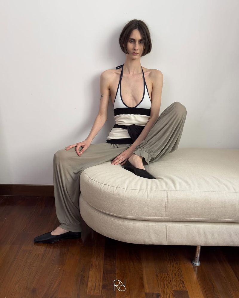 An adult woman is sitting on a beige couch indoors. She is wearing a halter top and trousers. There is a white wall in the background.