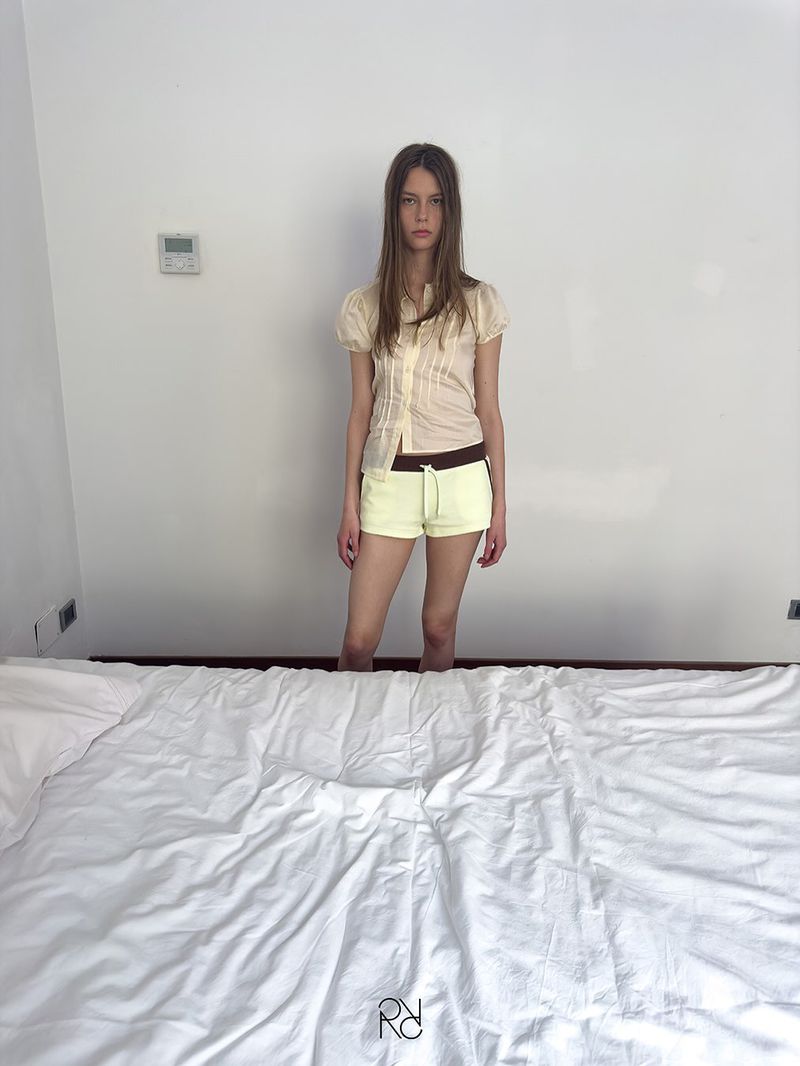 Young woman in cream top and yellow shorts stands by a made bed in an indoor setting with white walls, creating a simple fashion portrait.