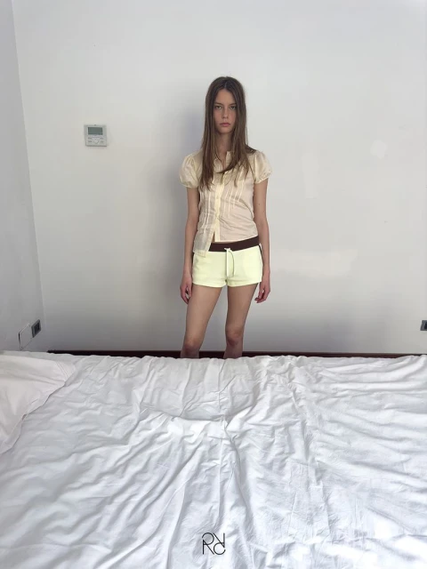 Young woman in cream top and yellow shorts stands by a made bed in an indoor setting with white walls, creating a simple fashion portrait.