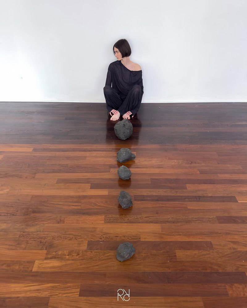 Woman sits with dark hair, wearing black clothing, aligned behind five scattered black rocks on dark wooden floor in front of a white wall.