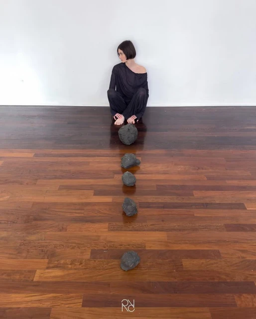 Woman sits with dark hair, wearing black clothing, aligned behind five scattered black rocks on dark wooden floor in front of a white wall.