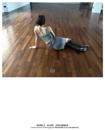 A slender woman with short hair sits on a wood floor in a gray dress and dark socks with back to camera in a fashion editorial.