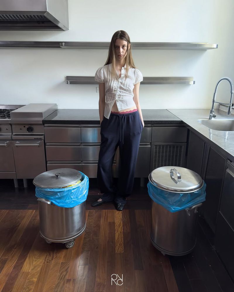 Young adult woman in white top and blue pants standing between two metal garbage cans inside of a modern kitchen space.