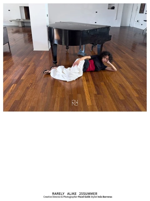 Adult female laying on a wood floor in front of a black grand piano. The room is well lit with sunlight and white walls.