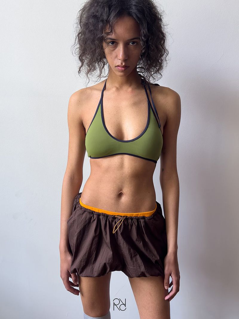 A young adult female poses in a halter top and bloomer shorts, indoors against a white wall, in a fashion or editorial setting.