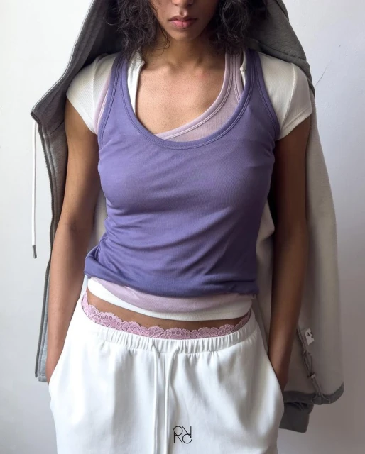 Woman wearing a purple tank top and white sweatpants poses in a casual indoor setting with a gray hooded sweatshirt.