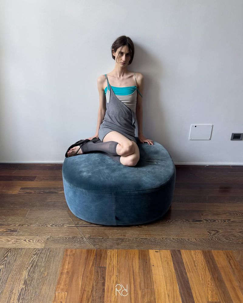 A young adult woman with dark hair sits on a blue ottoman in an indoor setting, modelling casual fashion in a relaxed pose against a white wall.