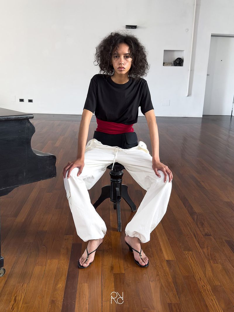 Young adult female sits on a stool in a studio, wearing a black t-shirt, red belt, white pants, and heels, with a piano in the background.