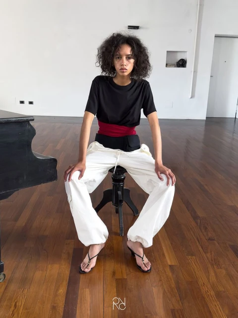 Young adult female sits on a stool in a studio, wearing a black t-shirt, red belt, white pants, and heels, with a piano in the background.