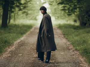 Alfie Nickerson walks along a tree-lined dirt path wearing a long dark overcoat and black boots, looking back over his shoulder.