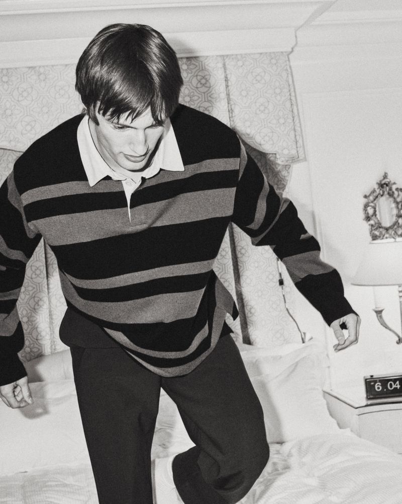 Model Thatcher Thornton wearing a dark striped rugby shirt and trousers while stepping on a white bed.