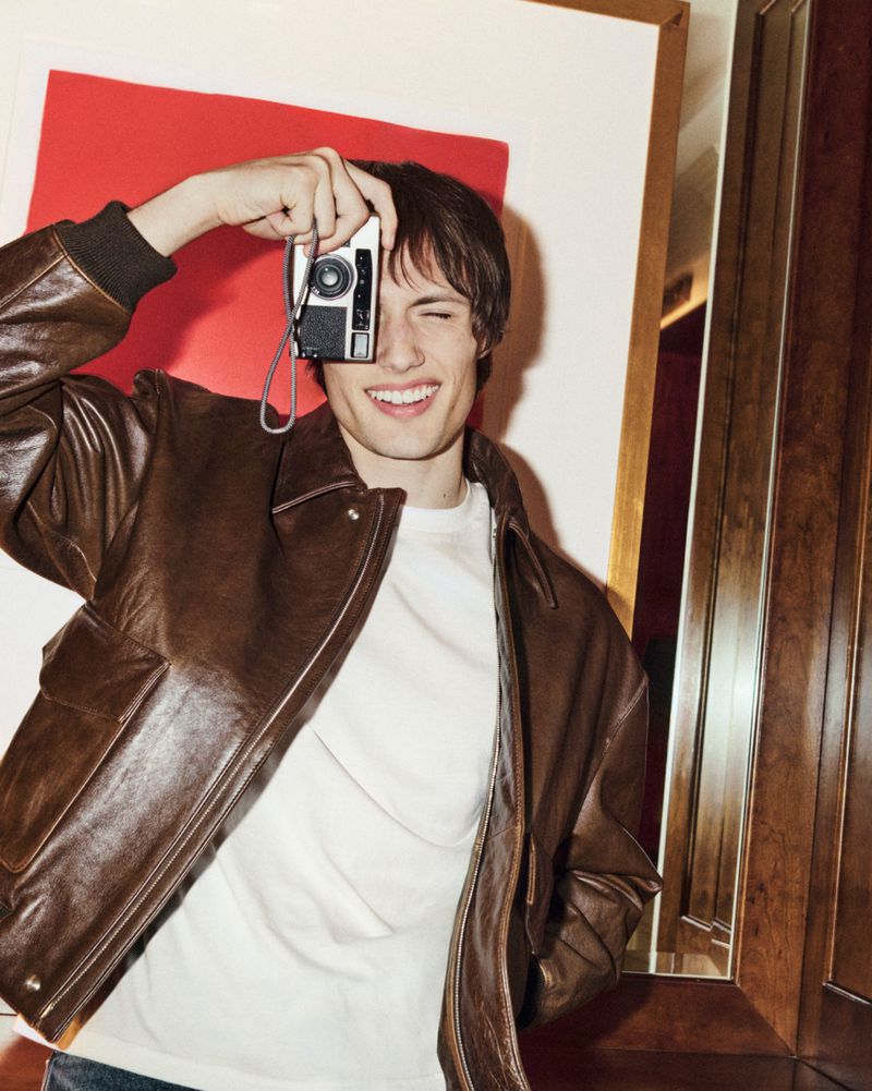 A young man in a brown leather bomber jacket smiles while holding a small vintage film camera up to his eye, indoor setting.