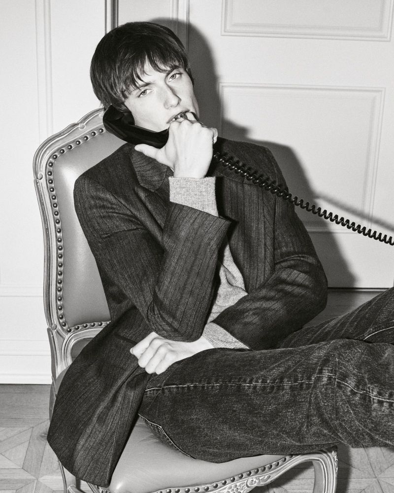 Model Thatcher Thornton sitting in an antique armchair holding a black corded telephone receiver in a pinstripe blazer and dark jeans.