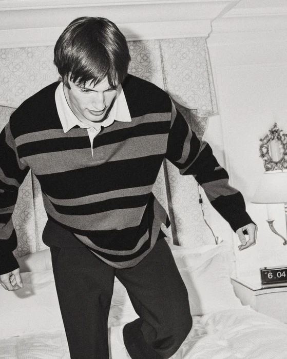 Model Thatcher Thornton wearing a dark striped rugby shirt and trousers while stepping on a white bed.