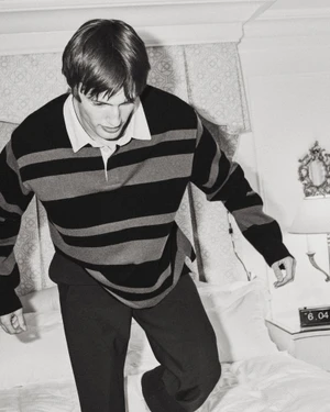 Model Thatcher Thornton wearing a dark striped rugby shirt and trousers while stepping on a white bed.