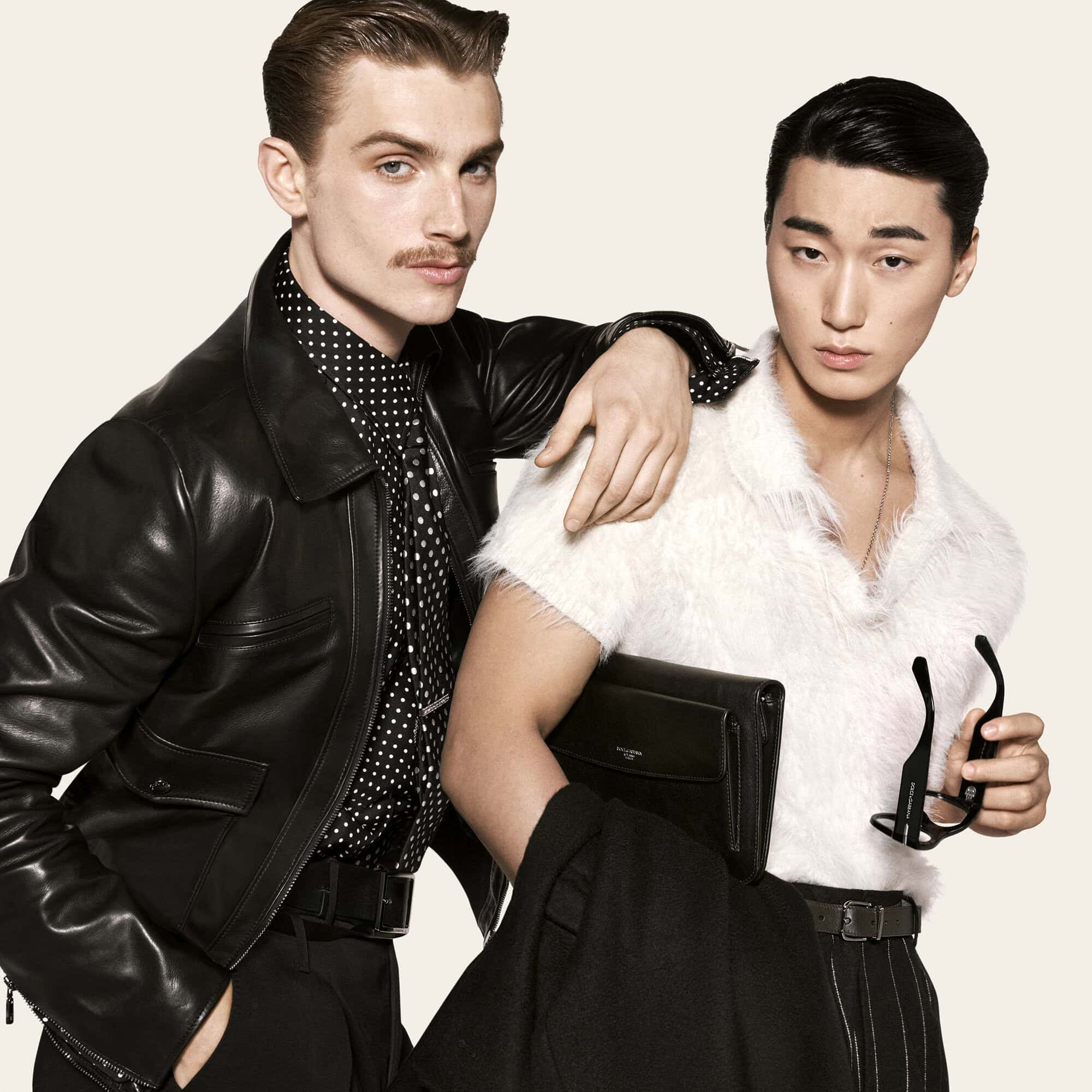 Two male models in a black leather jacket and fuzzy white polo shirt posing together against an off-white studio backdrop.