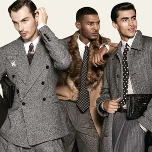 Three male models in tailored menswear, tweed suits, fur-trimmed coat, and ties posed against a clean light studio backdrop.