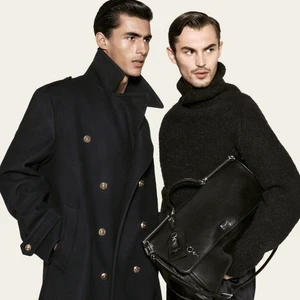 Two men posing in tailored black outerwear and a turtleneck sweater, with one holding a black leather briefcase against a pale backdrop.