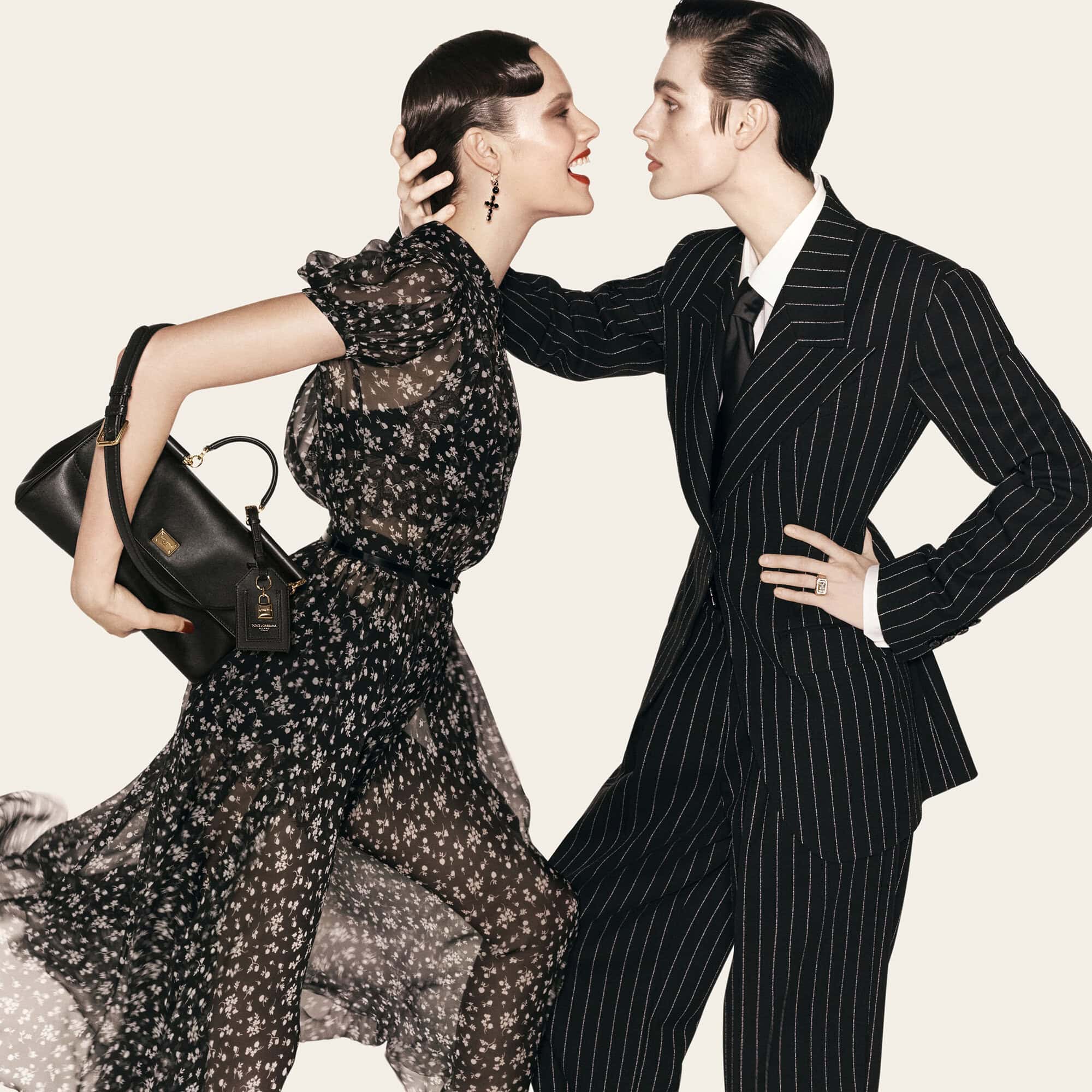 Two models in black outfits pose together, one wearing a floral dress holding a black handbag and the other in a pinstripe suit.