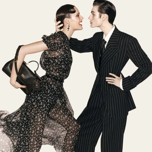 Two models in black outfits pose together, one wearing a floral dress holding a black handbag and the other in a pinstripe suit.