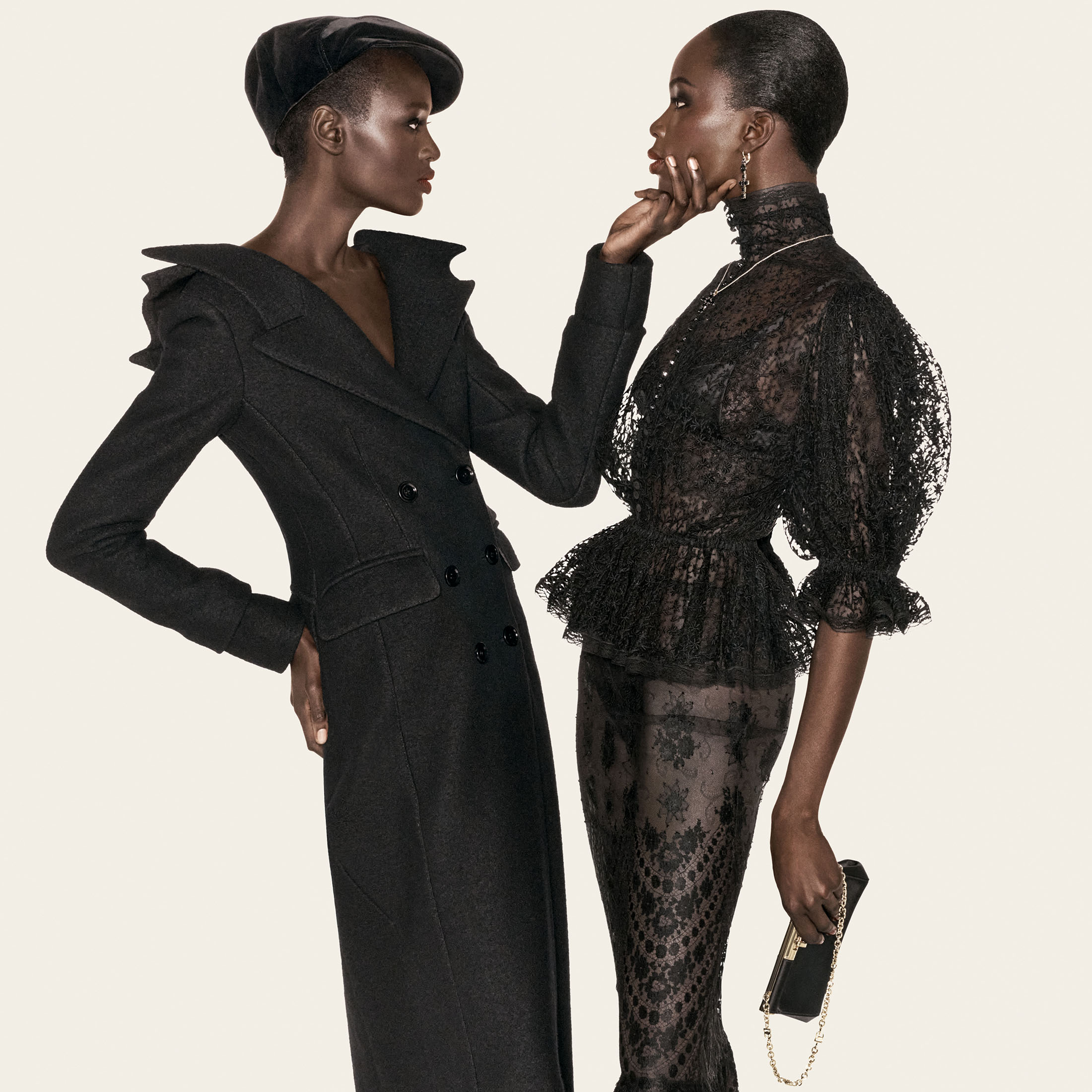 Two Black female models posing closely in all-black designer attire, one wearing a tailored coat and cap, the other in sheer black lace.