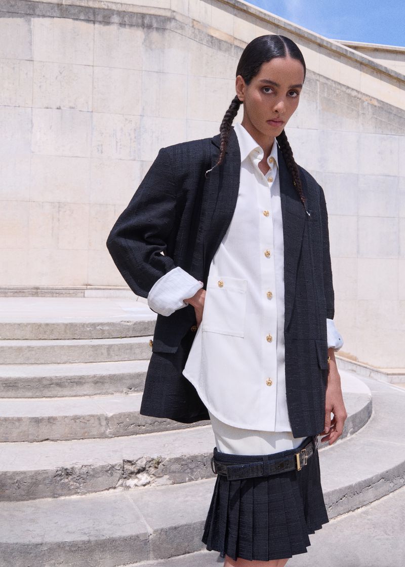 A woman with dark hair in two braids wears an oversized black blazer over a white shirt and a black pleated skirt, standing on stairs.