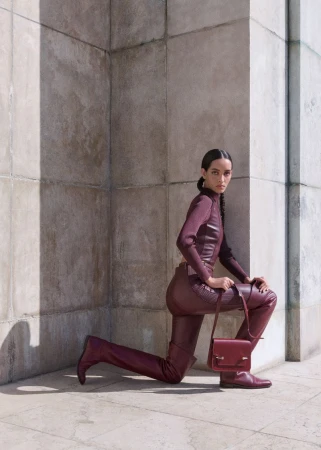 Model Eduarda Vieira poses kneeling in a monochrome burgundy leather outfit holding a matching handbag beside a stone column.