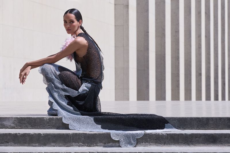 Model crouching on stone steps wearing a textured black gown with a trailing hem against tall architectural columns.