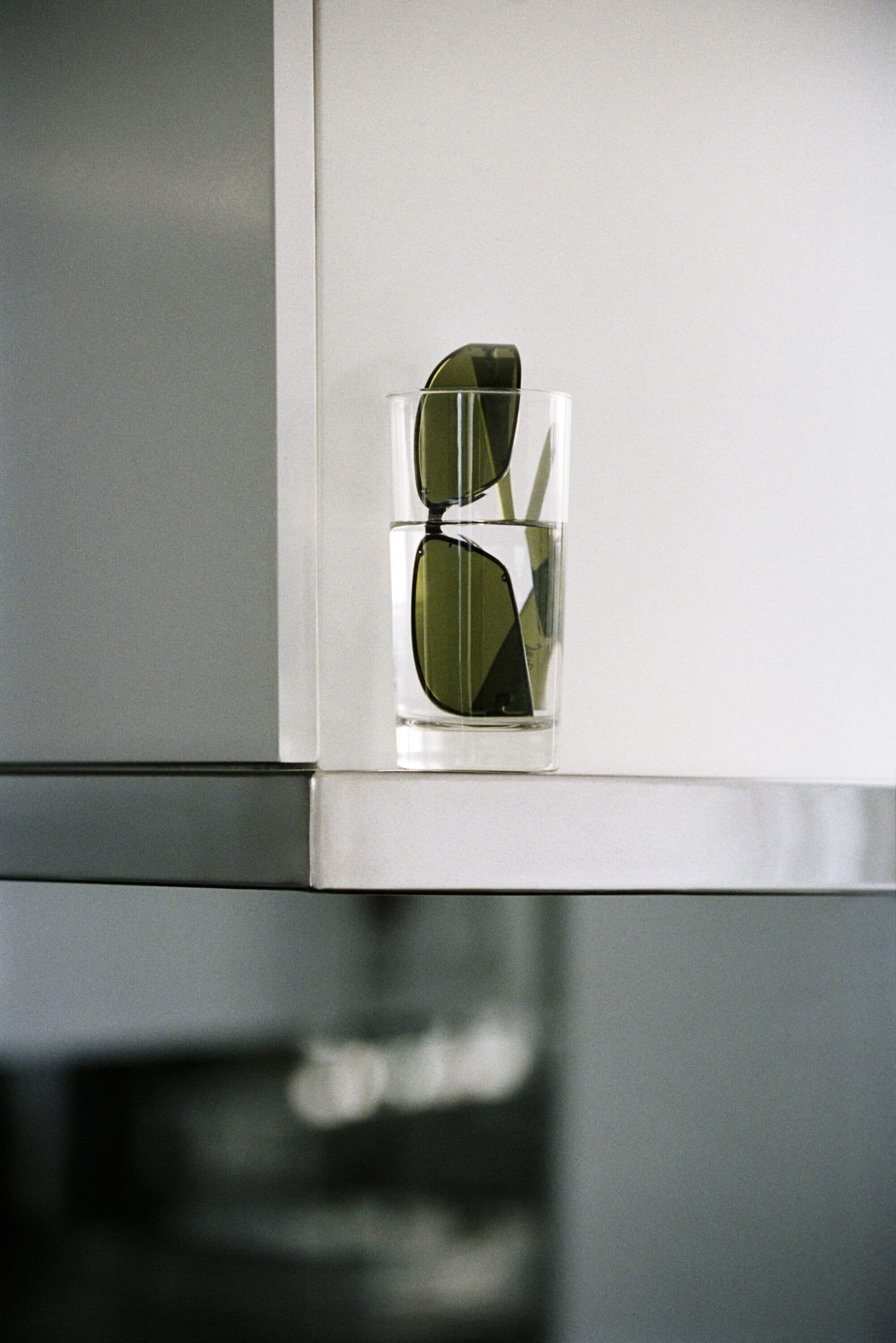 Dark green tinted sunglasses placed inside a clear glass tumbler of water on a sleek metallic-edged white shelf.
