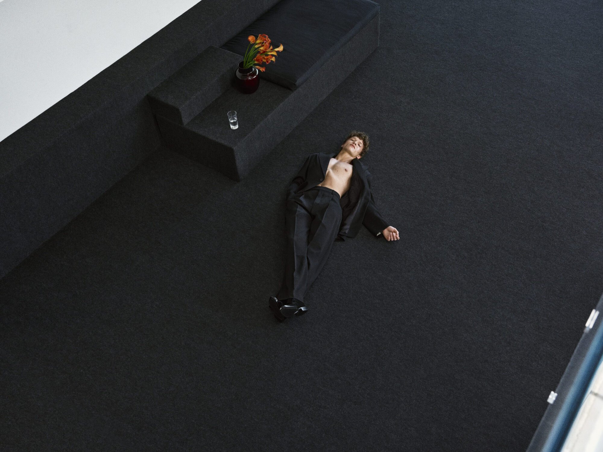 Connor Storrie lies on a black carpeted floor wearing an unbuttoned black suit jacket and trousers for Saint Laurent.
