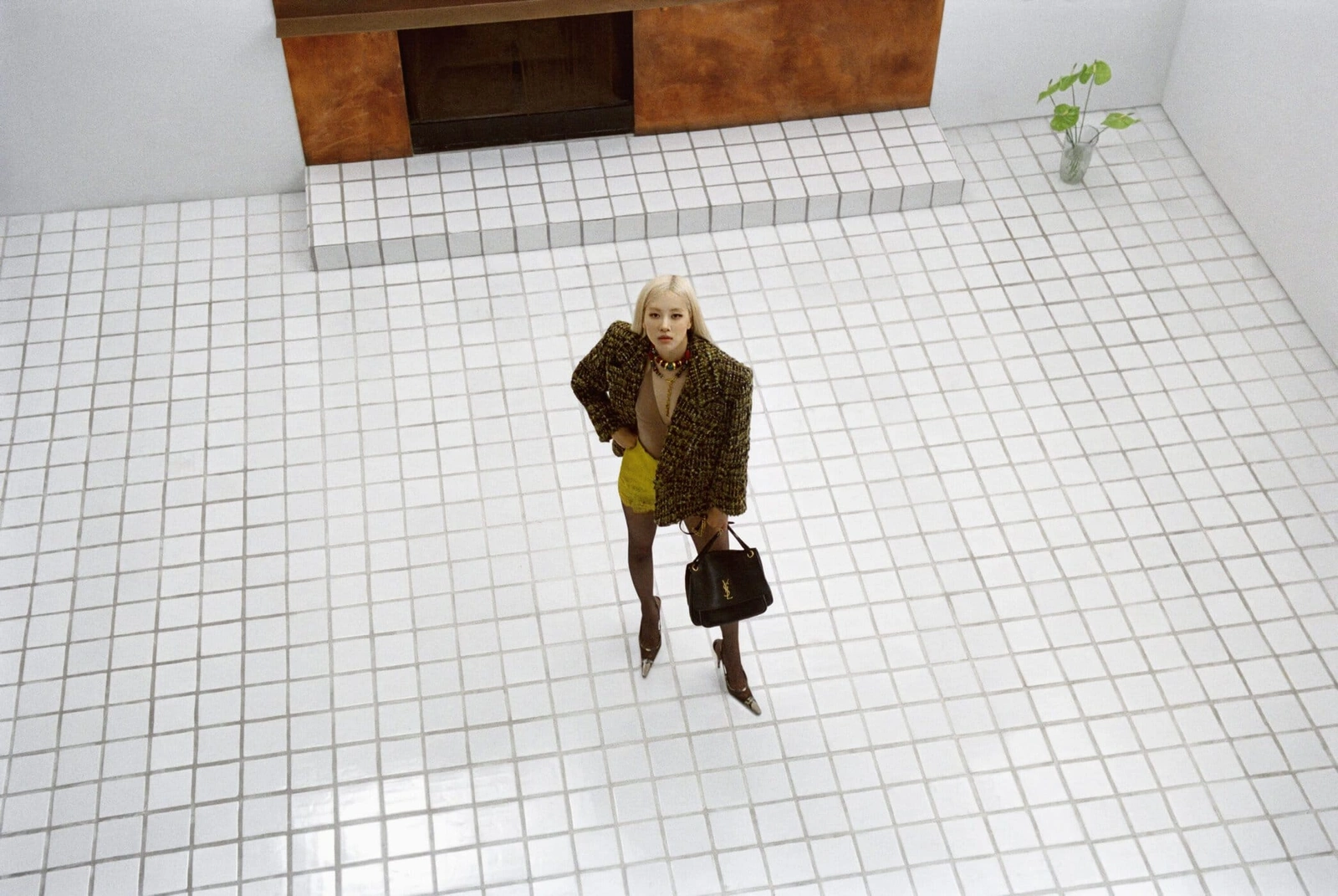 High-angle view of Rosé wearing an oversized brown tweed jacket, yellow shorts, and holding a black handbag on a white tiled floor.