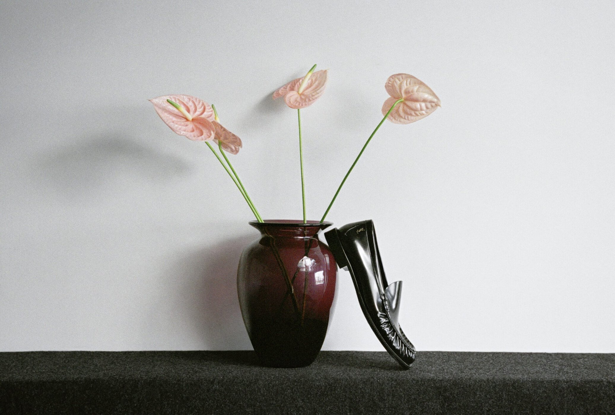 Black patent leather loafer propped against a dark burgundy vase filled with pink anthurium flowers on a dark surface.