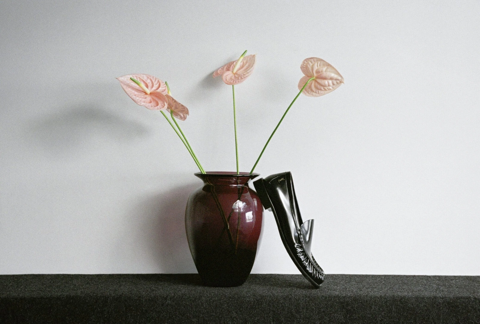 Black patent leather loafer propped against a dark burgundy vase filled with pink anthurium flowers on a dark surface.