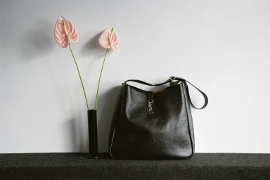 Black pebbled leather Saint Laurent shoulder bag next to two pink anthurium flowers in a black vase on a dark shelf.