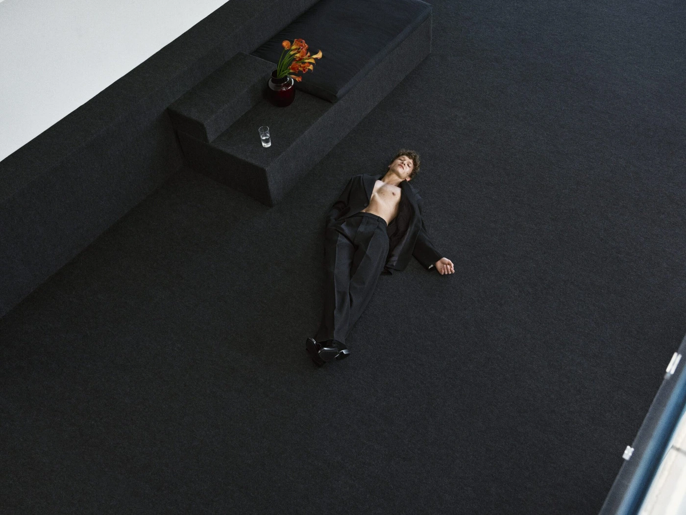 Connor Storrie lies on a black carpeted floor wearing an unbuttoned black suit jacket and trousers for Saint Laurent.