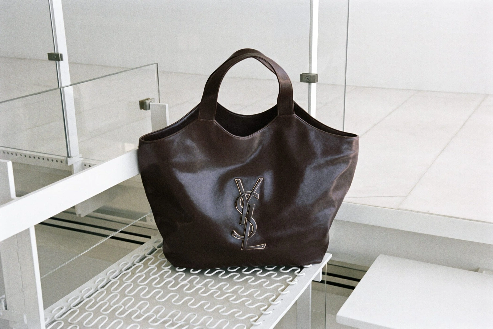 Dark brown leather Saint Laurent tote bag with metallic YSL monogram resting on a modern white mesh shelf.