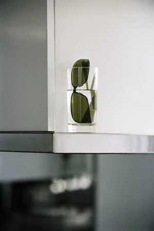 Dark green tinted sunglasses placed inside a clear glass tumbler of water on a sleek metallic-edged white shelf.