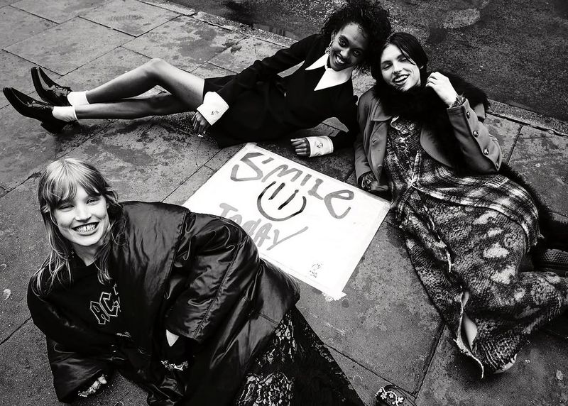 Three models smiling while lying on a paved city sidewalk around a handwritten Smile Today sign in a black and white editorial.