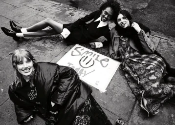 Three models smiling while lying on a paved city sidewalk around a handwritten Smile Today sign in a black and white editorial.