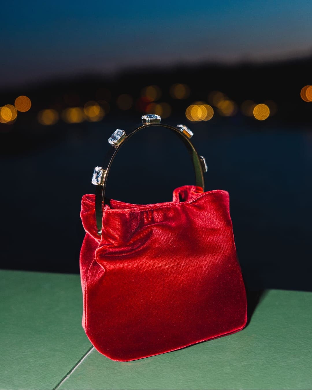 A small red velvet handbag with a gold-tone metal arch handle adorned with clear rectangular crystals, set against a blurred night waterfront.