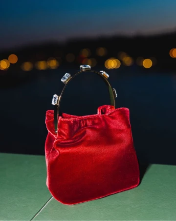 Red velvet handbag with a crystal-embellished curved handle resting on a green ledge against blurred night city lights.