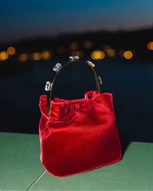 Red velvet handbag with a crystal-embellished curved handle resting on a green ledge against blurred night city lights.