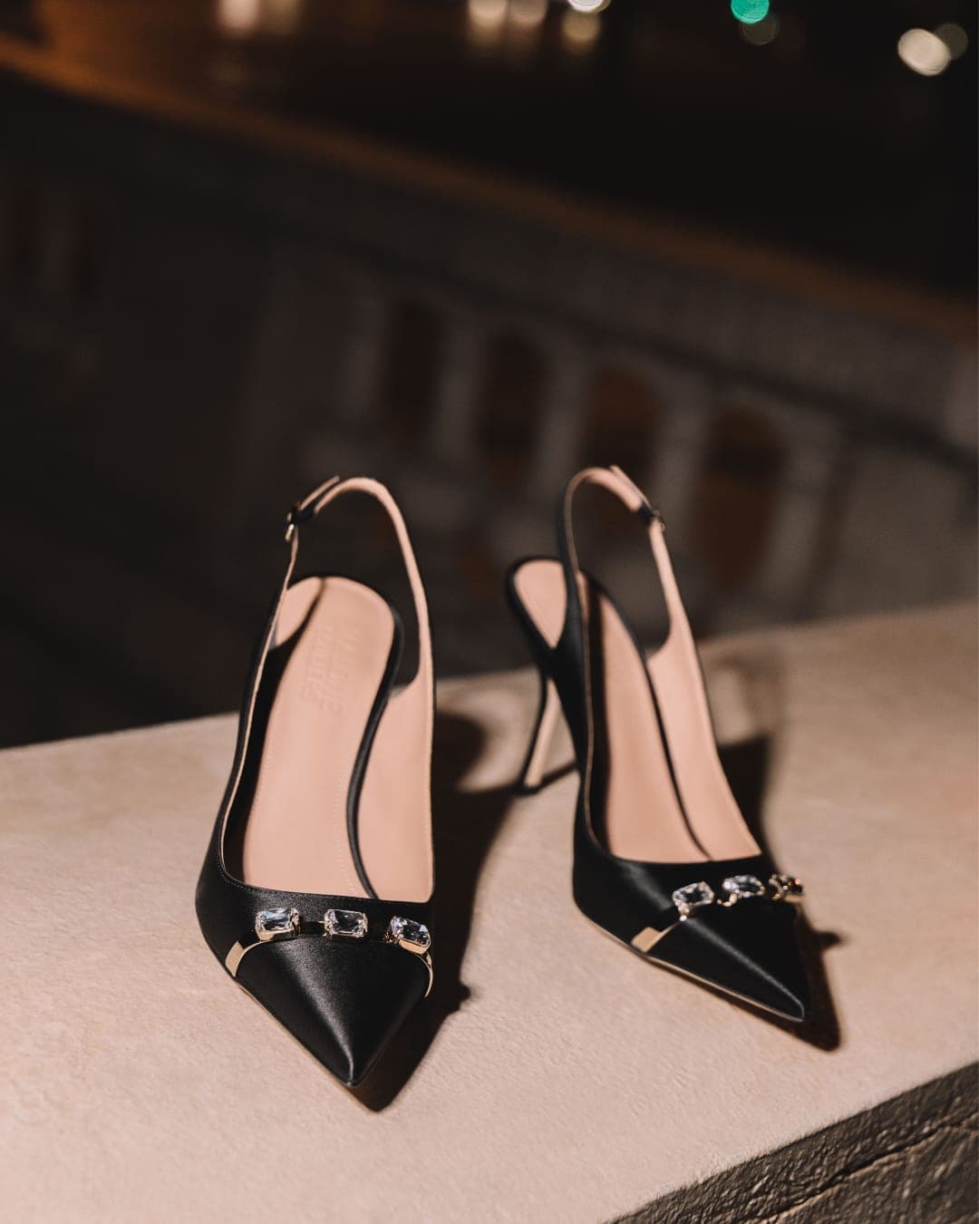 Pair of black satin pointed-toe slingback heels with crystal embellishments resting on a stone ledge at night.