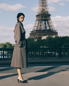 Model Kirandeep Chahal wearing a taupe trench coat and black mule heels standing on a bridge in front of the Eiffel Tower.