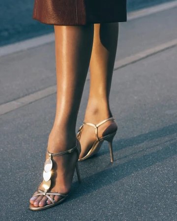 Close-up of a model wearing metallic gold strappy stiletto sandals with disc details on an asphalt road.